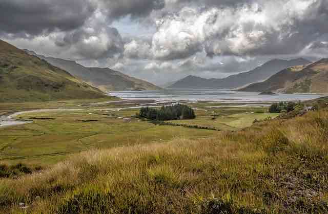 view of a wide green valley and a lock surrounded by mountains and clouds in the sky