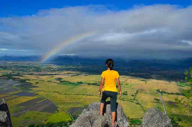 woman standing on a rock overlooking a green valley with a rainbow